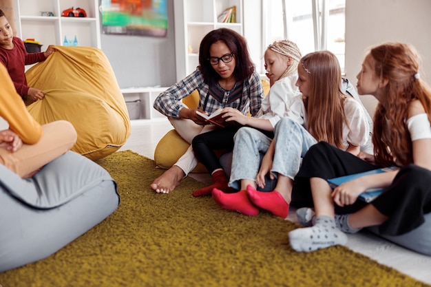 A teacher reads a book to a small group of attentive preschool children sitting on a colorful rug. The children are diverse and the environment is warm and inviting.