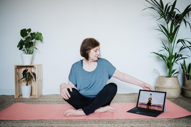 A remote worker is participating in a virtual wellness session that is focused on stretching and mindfulness. The worker is relaxed and engaged, and the instructor is shown on a laptop screen in a well-lit and comfortable setting.
